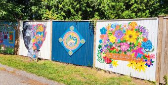 photo of 3 large fence panels, each painted differently. one is a human face that's painted half in black and white and half in vibrant colors, next to that is a peacock blue panel with a diamond-shaped panel in its center. on that diamond is a painting of a peacock in a mosaic style. the last panel is covered almost entirely by a painting of a brilliant bouquet of flowers with the phrase the most beautiful flower underneath.