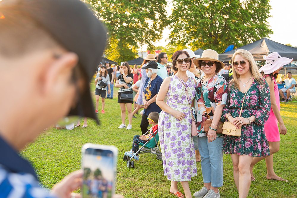 photo of a person holding a phone to take a picture of 3 smiling friends attending an outdoor festival