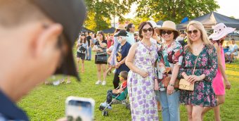 photo of a person holding a phone to take a picture of 3 smiling friends attending an outdoor festival