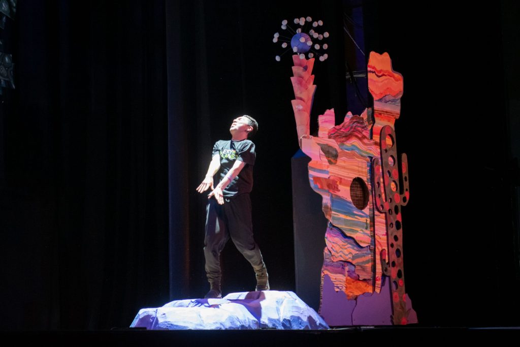 production still of an actor standing under a spotlight making an anguished face. Behind them in the shadows is an abstract set piece that's multi-colored and contains elements that resemble motifs of the Southwestern United States.