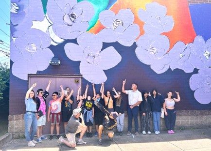 Photo of 12 students and a teacher pointing upwards at the colorful mural behind them, consisting of purple flowers on a colorful dark blue and orange background. Two teaching artists kneel in the ground in front of them and also point up.