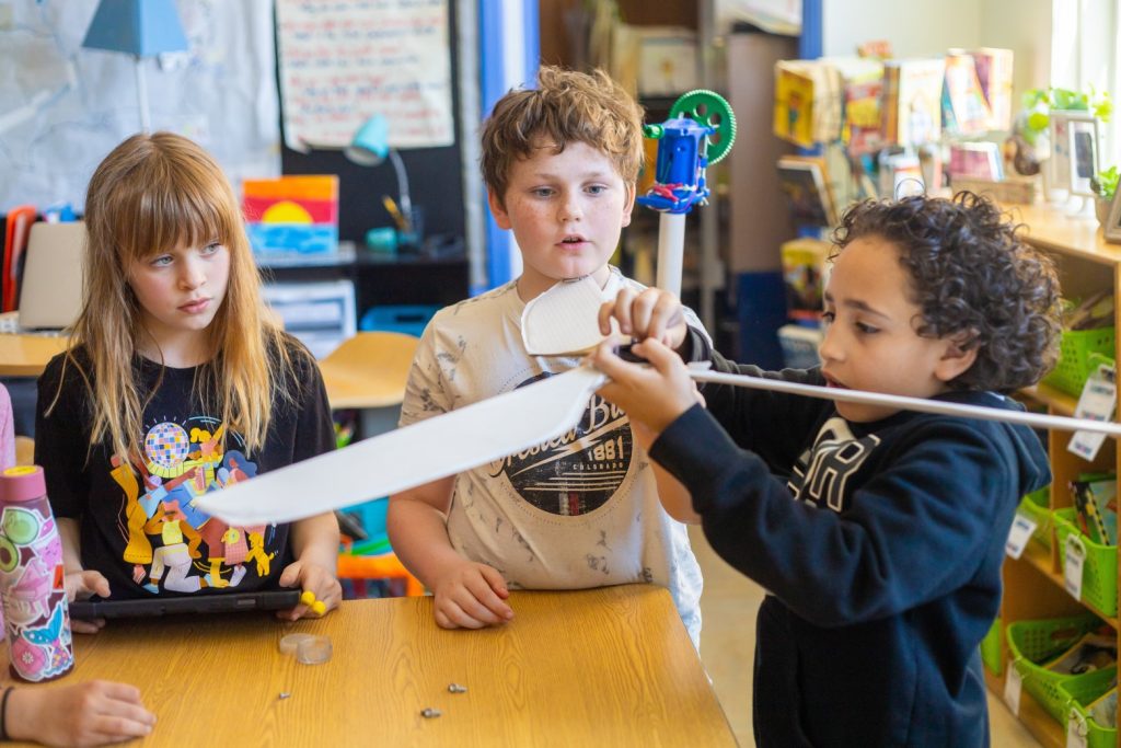 Photo of 2 fourth grade students looking on intently at a third student holding the self-constructed wings of a wind turbine.