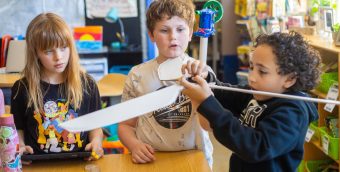 Photo of 2 fourth grade students looking on intently at a third student holding the self-constructed wings of a wind turbine.