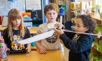 Photo of 2 fourth grade students looking on intently at a third student holding the self-constructed wings of a wind turbine.