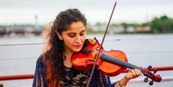 Photograph of a Latina woman deep in the experience of playing her violin, with the Boston Harbor behind her in the distance.