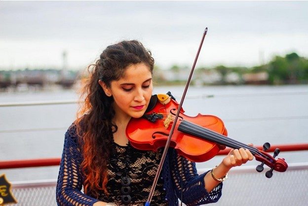 Photograph of a Latina woman deep in the experience of playing her violin, with the Boston Harbor behind her in the distance.