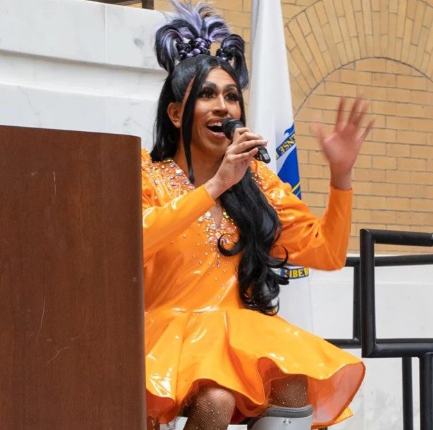 A drag artist wearing a vibrant orange dress, singing passionately into a microphone.