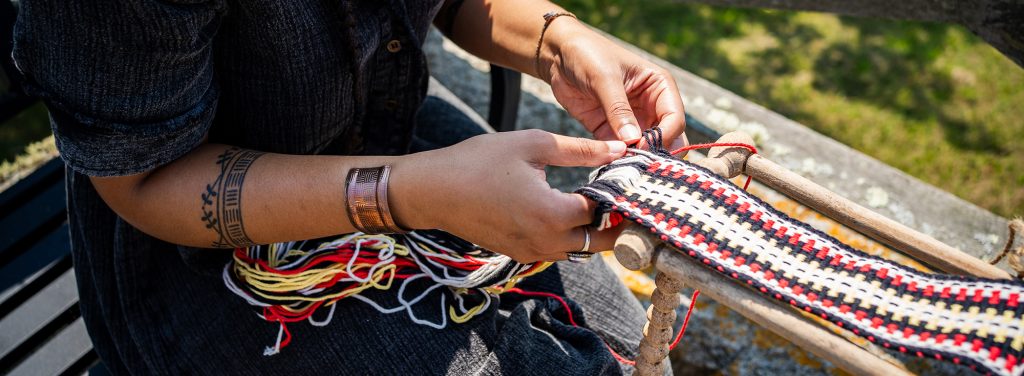 Photo detail of a pair of hands weaving many strands of white, red, yellow, and black into a traditional twined sash.