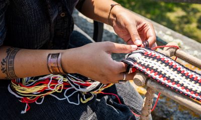 Photo detail of a pair of hands weaving many strands of white, red, yellow, and black into a traditional twined sash.