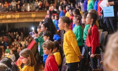 photo taken from the side of a few rows of children singing. They are wearing long sleeve t-shirts in solid primary colors.