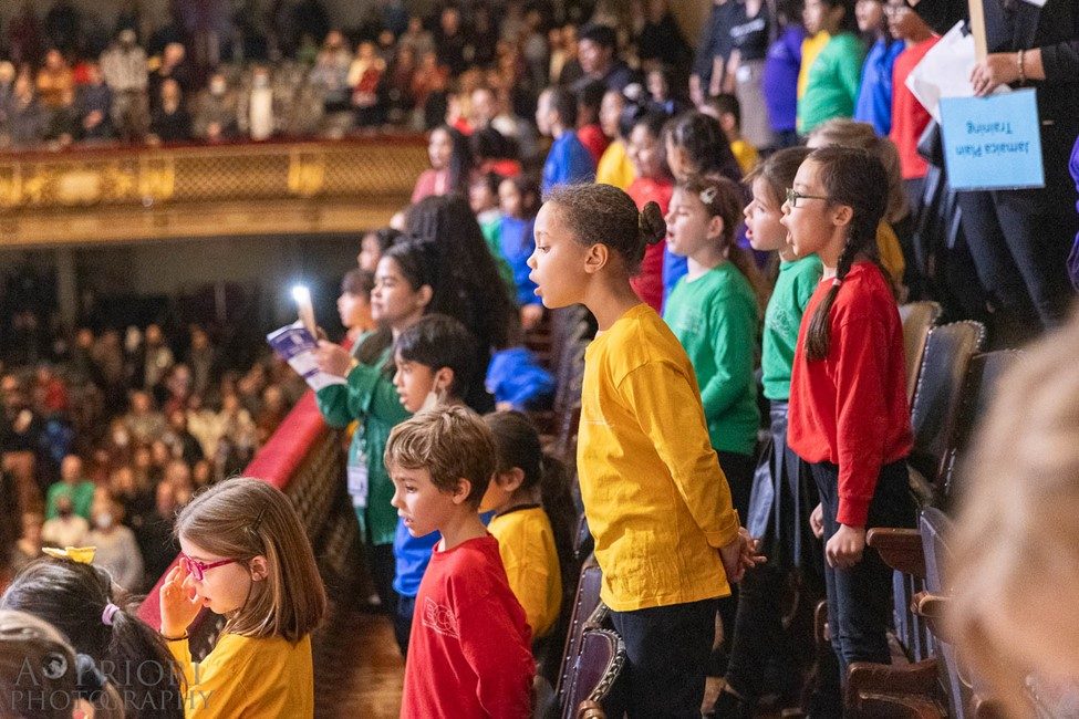 photo taken from the side of a few rows of children singing. They are wearing long sleeve t-shirts in solid primary colors.