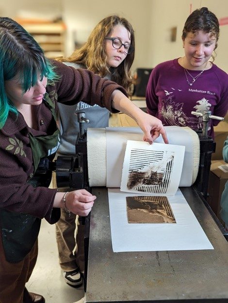 photo of 2 young artists looking on while a 3rd young artist carefully pulls a freshly printed art piece off the press.