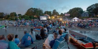 photo of hundreds of people sitting on the beach at dusk watching a film projected on a screen.