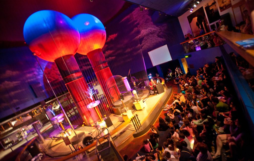 photo of Museum of Science's Theater of Electricity, featuring a demonstration of the world's largest air-insulated Van de Graaff generators to a room full of on-lookers. 