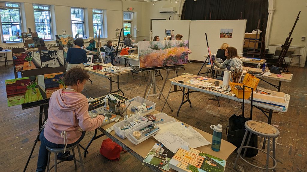 photo of a painting studio with numerous people seated painting canvases, tables are covered with materials and brushes.