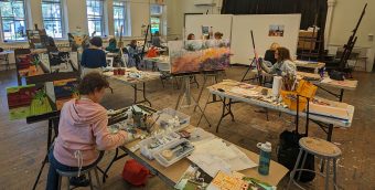 photo of a painting studio with numerous people painting canvases, tables are covered with materials and brushes.