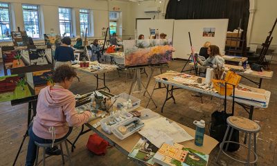 photo of a painting studio with numerous people painting canvases, tables are covered with materials and brushes.