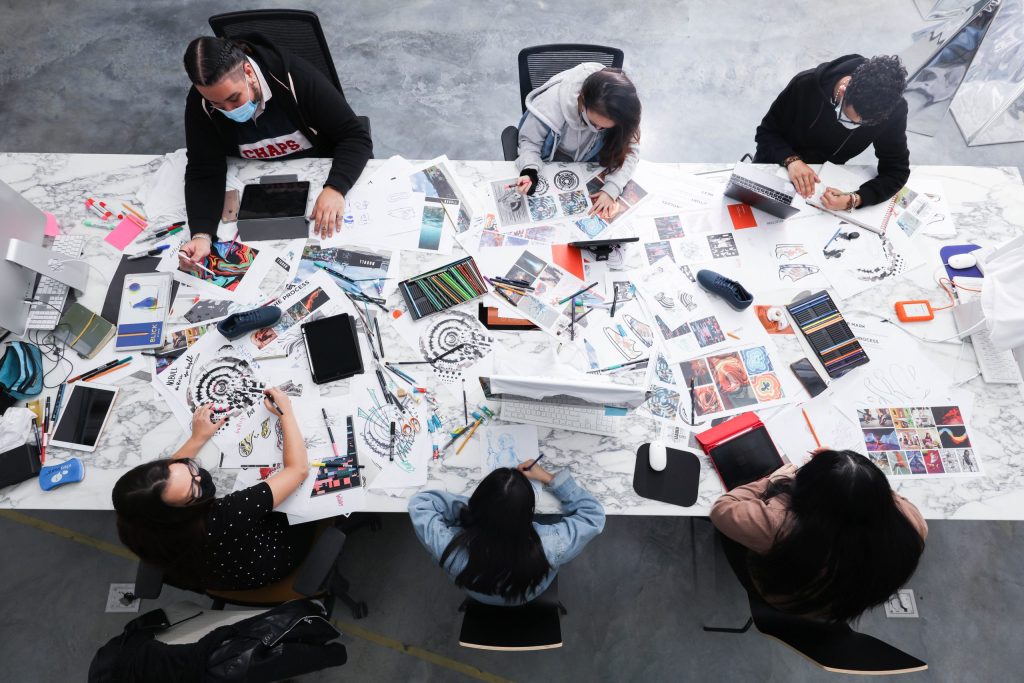 photo looking down at a large table covered in print outs and mock ups. 6 young people sitting in black office chairs work with the papers strewn about.