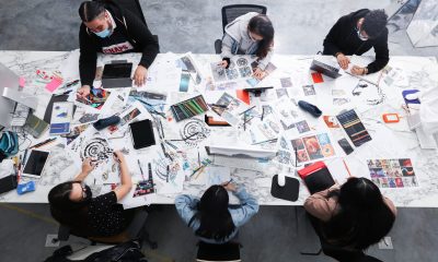 photo looking down at a large table covered in print outs and mock ups. 6 young people sitting in black office chairs work with the papers strewn about.