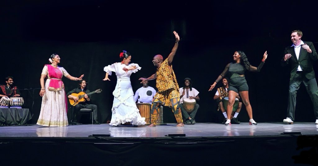 photo of a dance performance. 4 musicians play seated in the background while a half dozen dancers wearing outfits depicting their individual dance genre are in a line in the foreground.