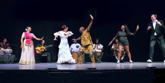 photo of a dance performance. 4 musicians play seated in the background while a half dozen dancers wearing outfits depicting their individual dance genre are in a line in the foreground.