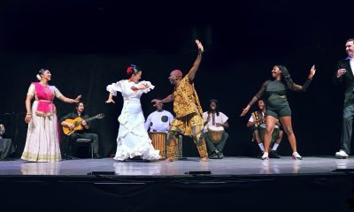 photo of a dance performance. 4 musicians play seated in the background while a half dozen dancers wearing outfits depicting their individual dance genre are in a line in the foreground.