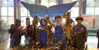 Five teen Apprentices standing behind a large pile of seaweed, smiling and holding up seaweed with a decorative blue whale tail as a backdrop.