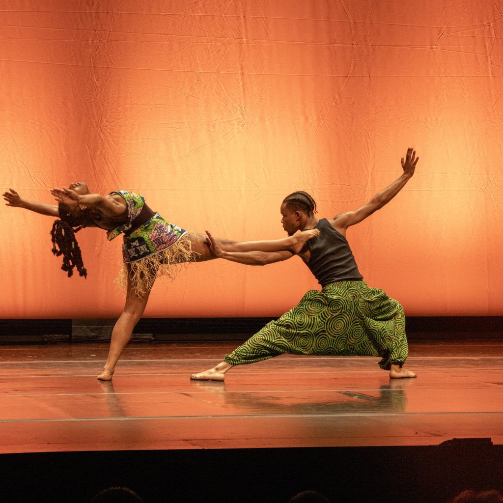 Two young African American dancers, one with short, cropped hair and one with long hair pose dramatically on stage. A dancer in green pants holds the leg of the other dancer, who is balancing on one leg while leaning backwards elegantly, parallel to the ground. Both dancers have arms outstretched against an orange background.