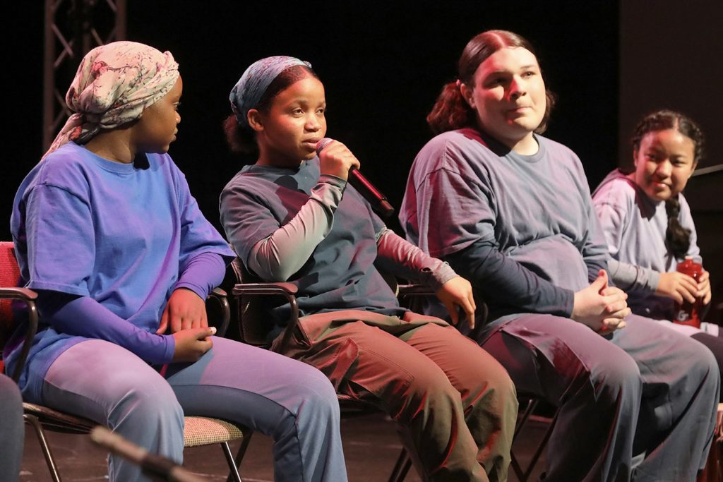 An African American teen is speaking and holding a microphone while three other teens of various races wait their turn to speaking in a post-show dialogue. 