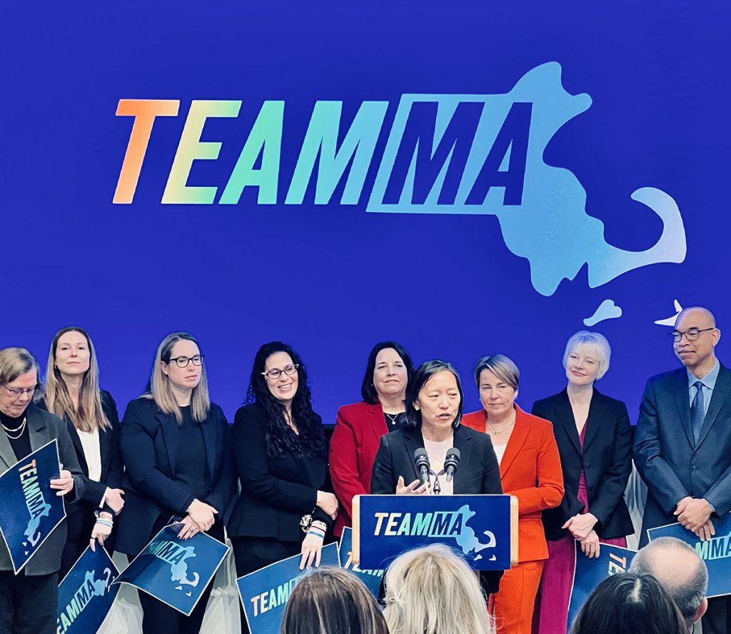 group photo of Governor Healey and her cabinet members. Yvonne Hao is speaking at a podium.