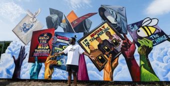 Michael Aghohawa stands before his mural. The mural shows blue, purple, yellow, and green hands holding and throwing up graduation caps in front of a bright sky. The graduation caps have text and drawings on them, including “I hold the keys to the future” in a key hole, “I’m history in the making” with three black women holding up awards, and “I will BEE the change” with an image of a bee.