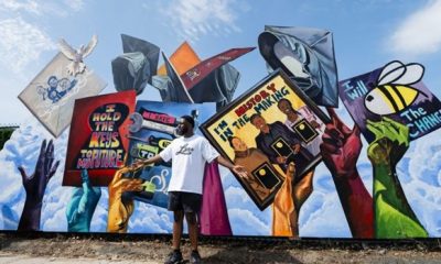 Michael Aghohawa stands before his mural. The mural shows blue, purple, yellow, and green hands holding and throwing up graduation caps in front of a bright sky. The graduation caps have text and drawings on them, including “I hold the keys to the future” in a key hole, “I’m history in the making” with three black women holding up awards, and “I will BEE the change” with an image of a bee.