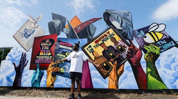 Michael Aghohawa stands before his mural. The mural shows blue, purple, yellow, and green hands holding and throwing up graduation caps in front of a bright sky. The graduation caps have text and drawings on them, including “I hold the keys to the future” in a key hole, “I’m history in the making” with three black women holding up awards, and “I will BEE the change” with an image of a bee.