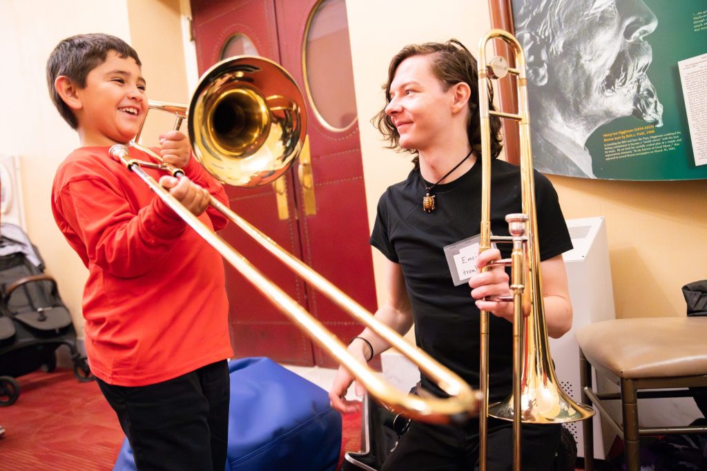 photo of a young person trying out a trombone. A seated adult musician also holding a trombone is encouraging him.