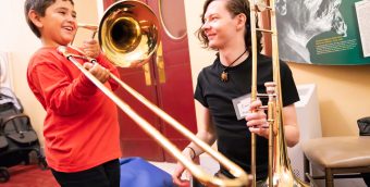 photo of a young person trying out a trombone. A seated adult musician also holding a trombone is encouraging him.