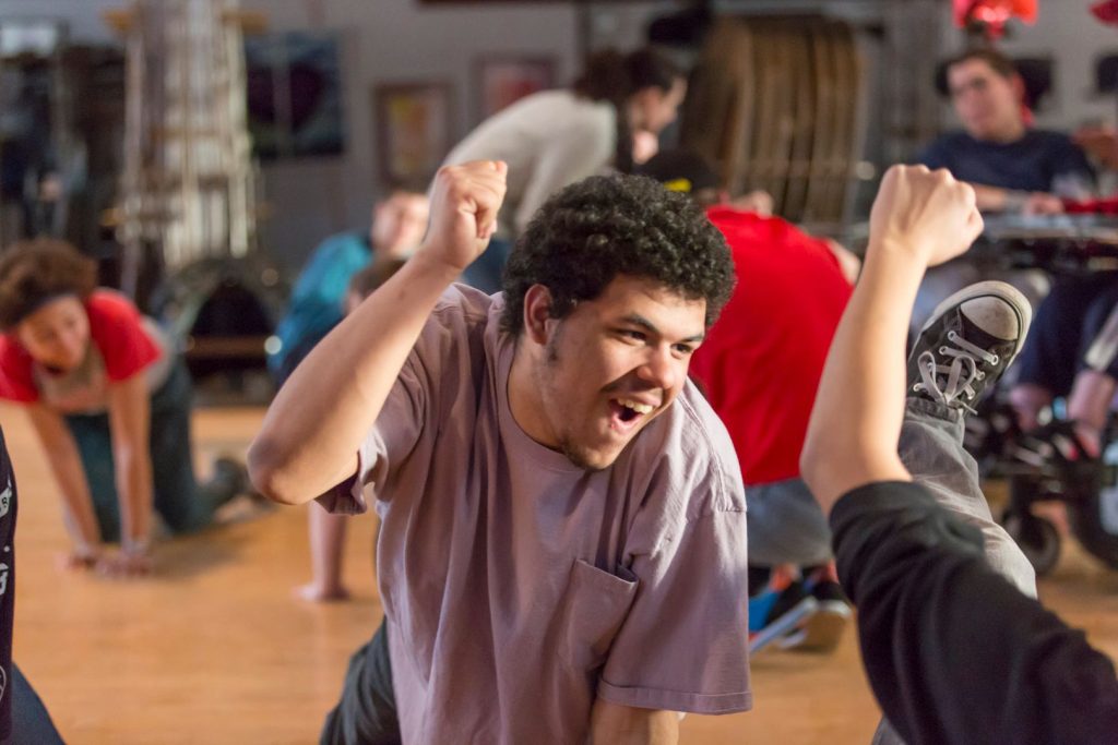 photo from a theater rehearsal. In the foreground a young man is on his hands and knees, his right hand raised in a triumphant fist, he's looking up and his face is beaming. In the background are other actors also on their hands and knees.