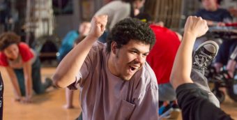 photo from a theater rehearsal. In the foreground a young man is on his hands and knees, his right hand raised in a triumphant fist, he's looking up and his face is beaming. In the background are other actors also on their hands and knees.