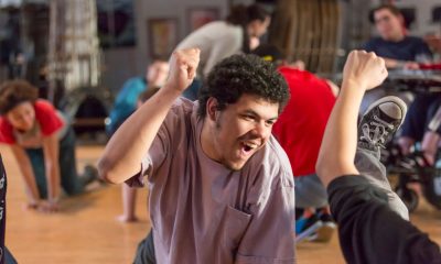 photo from a theater rehearsal. In the foreground a young man is on his hands and knees, his right hand raised in a triumphant fist, he's looking up and his face is beaming. In the background are other actors also on their hands and knees.