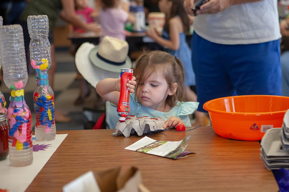 photo of a child with a very focused look holding a red bingo marker upside down and coloring the peaks of an upside down egg carton. In the background other young people are seated at tables working on other arts projects.