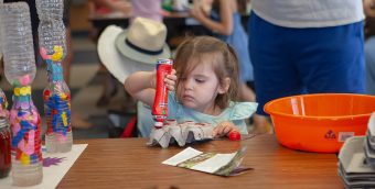 photo of a child with a very focused look holding a red bingo marker upside down and coloring the peaks of an upside down egg carton. In the background other young people are seated at tables working on other arts projects.