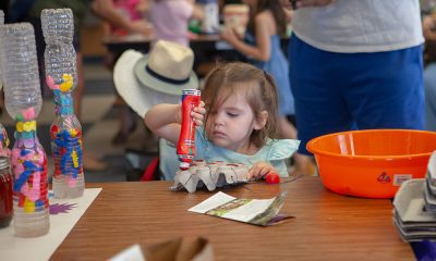 photo of a child with a very focused look holding a red bingo marker upside down and coloring the peaks of an upside down egg carton. In the background other young people are seated at tables working on other arts projects.