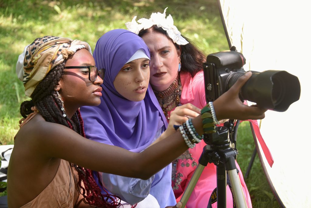 Two teens and a teaching artist, all of different races and colorful attired, intently look through a large camera with a telephoto lens.