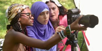 Two teens and a teaching artist, all of different races and colorful attired, intently look through a large camera with a telephoto lens.