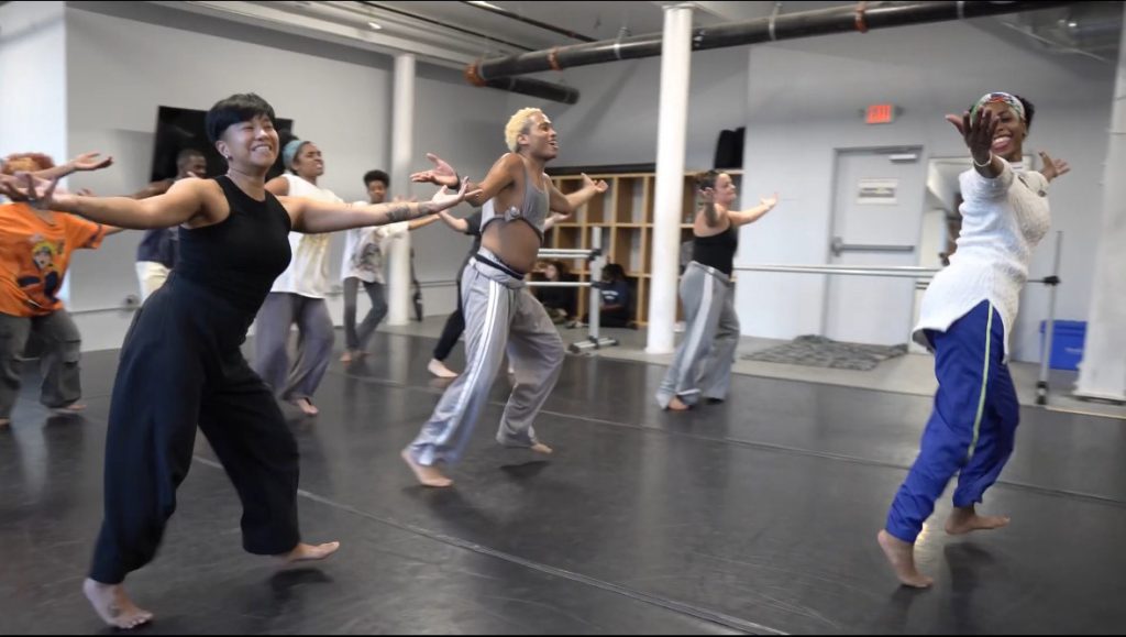 photo of about a dozen dancers having a rehearsal, they are wearing lose fitting athletic wear and have their arms extended, palms up, at their sides.