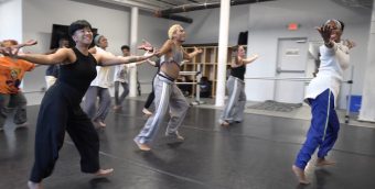 photo of about a dozen dancers having a rehearsal, they are wearing lose fitting athletic wear and have their arms extended, palms up, at their sides.