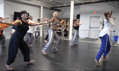 photo of about a dozen dancers having a rehearsal, they are wearing lose fitting athletic wear and have their arms extended, palms up, at their sides.