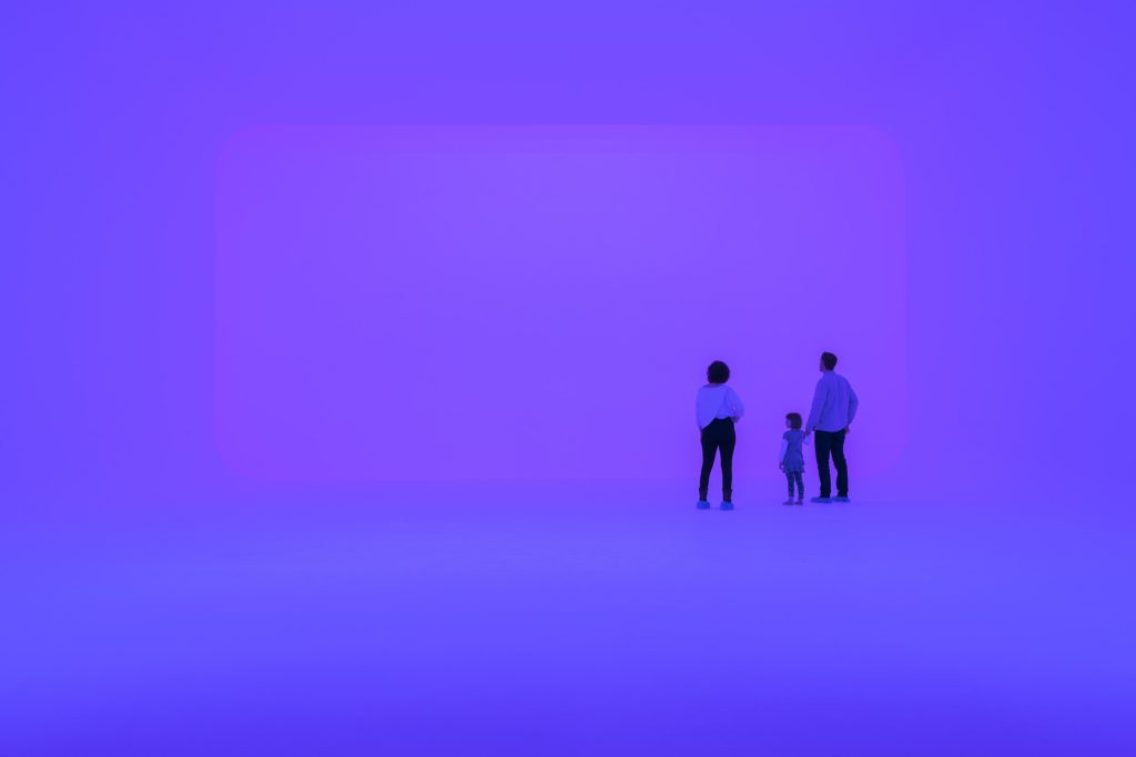 photo of 3 people standing in an immersive light experience, the space is glowing a purple-ish blue on all surfaces, evoking an infinite feeling of space. They are facing a wall with a large rectangular shape that is glowing a slightly different shade of purple from the rest of the space.