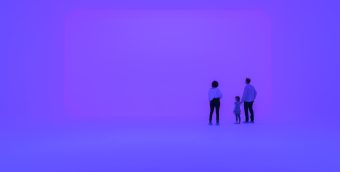 photo of 3 people standing in an immersive light experience, the space is glowing a purple-ish blue on all surfaces, evoking an infinite feeling of space. They are facing a wall with a large rectangular shape that is glowing a slightly different shade of purple from the rest of the space.