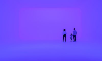 photo of 3 people standing in an immersive light experience, the space is glowing a purple-ish blue on all surfaces, evoking an infinite feeling of space. They are facing a wall with a large rectangular shape that is glowing a slightly different shade of purple from the rest of the space.