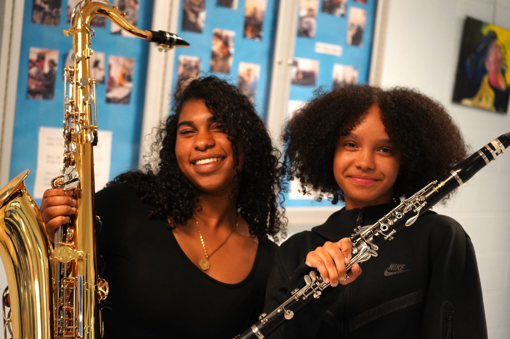 Two young women with black curly hair hold their instruments up and smile. One has a saxophone, the other a clarinet. 